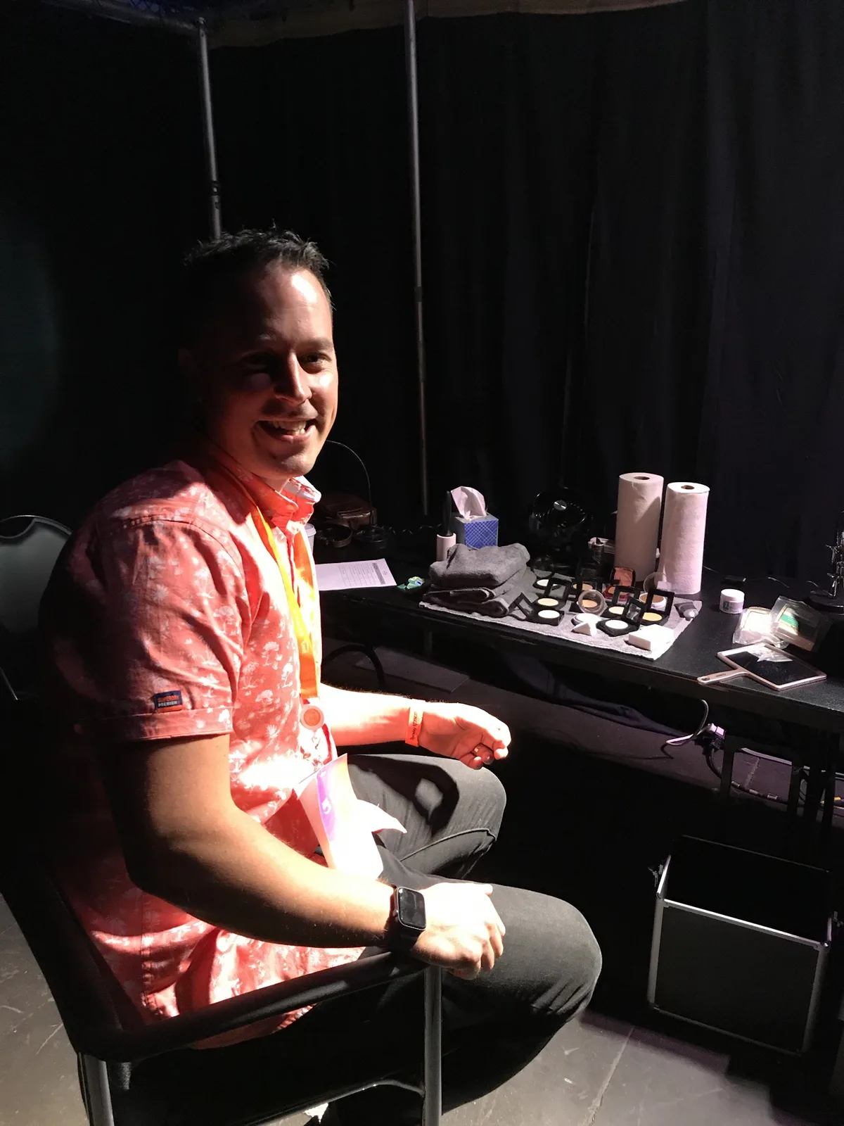 Martin in a pink floral shirt, grinning in a make-up chair backstage, with a table of make-up and paper towels behind him.