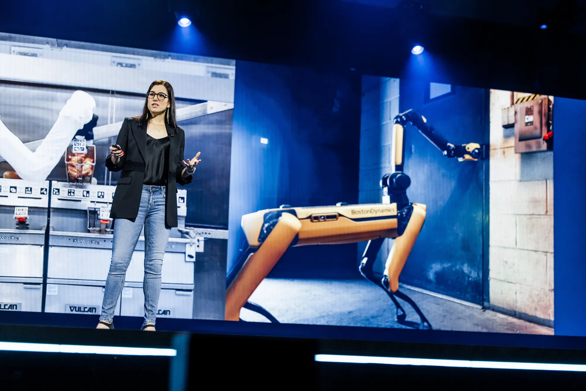 Roni Sole on stage beside footage of a robot dog in a warehouse