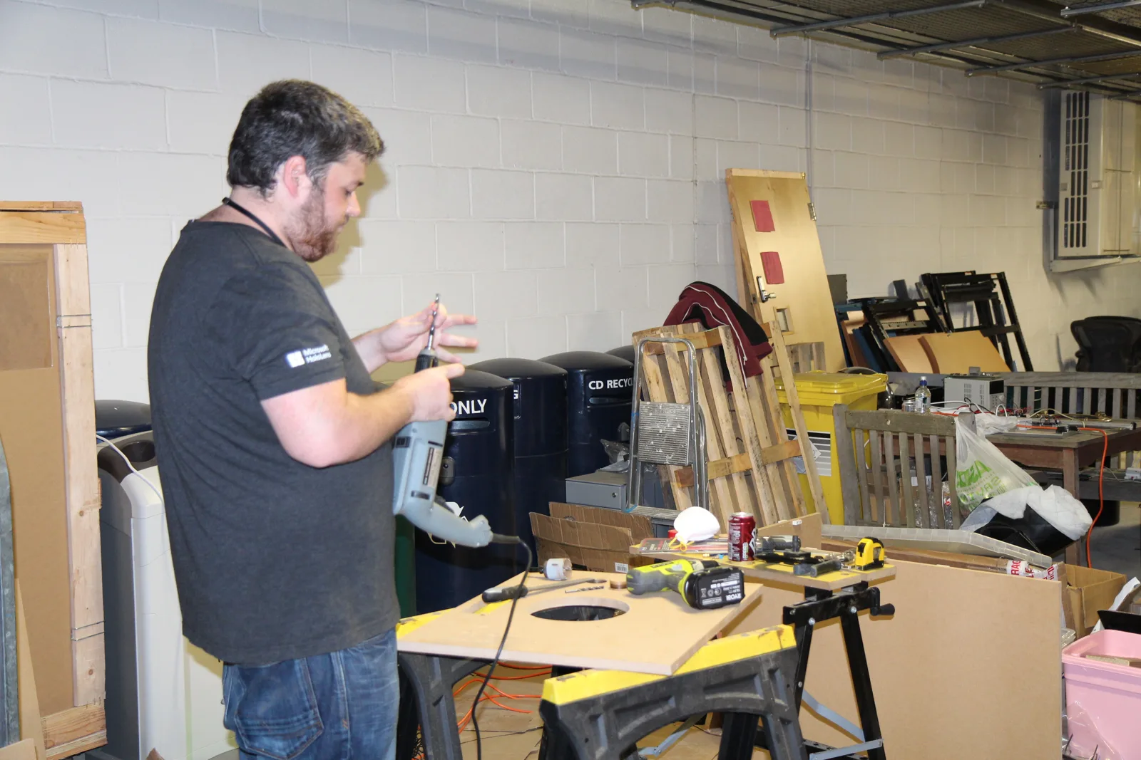 A builder at a workbench fitting parts into a wooden panel with a round hole cut for a dial.