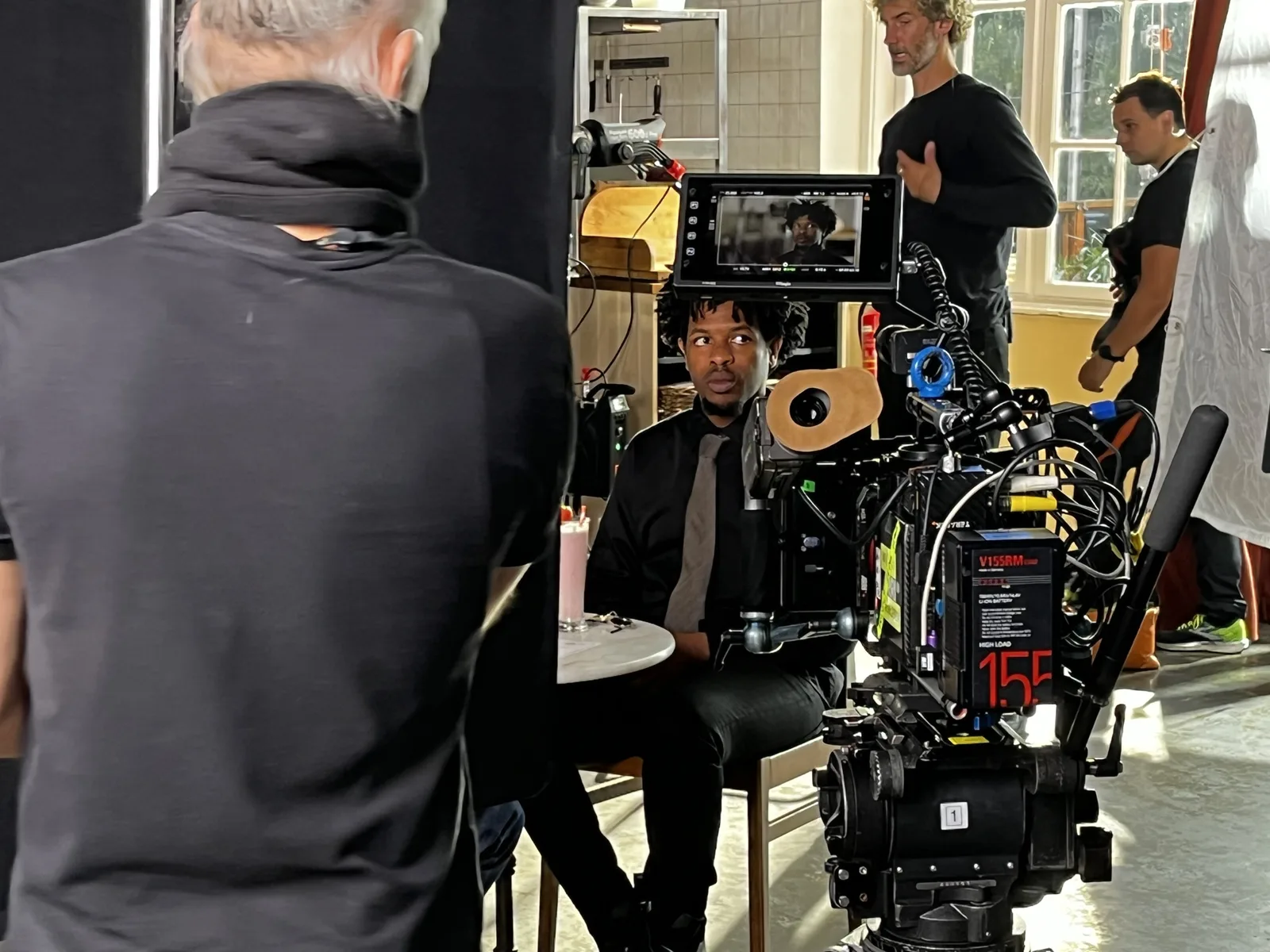 Terrance Lamonte Jr., in a black shirt and tie, sitting at a café table behind the camera rig between takes.
