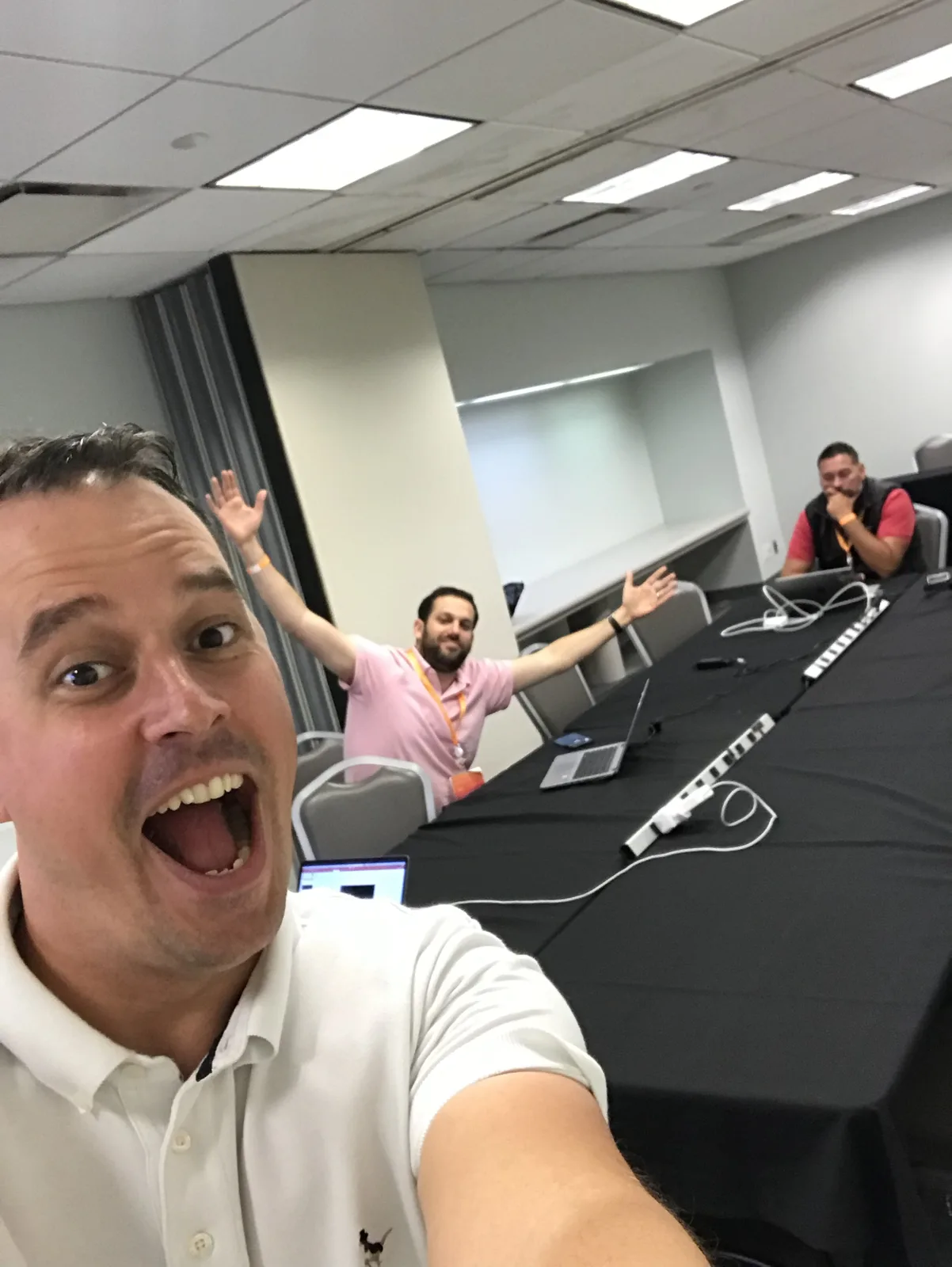 A selfie of Martin pulling a delighted face in the demo prep room, with two colleagues behind him at a long table of laptops and cables.