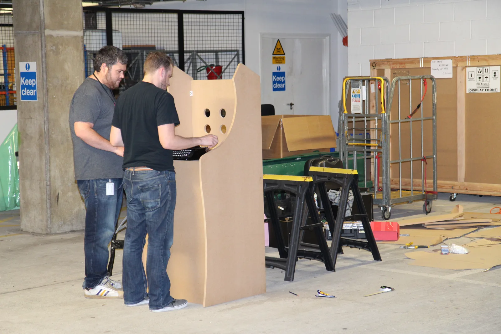 Two of the builders studying a full-size cardboard mock-up of the cabinet, with workbenches and offcuts around them.