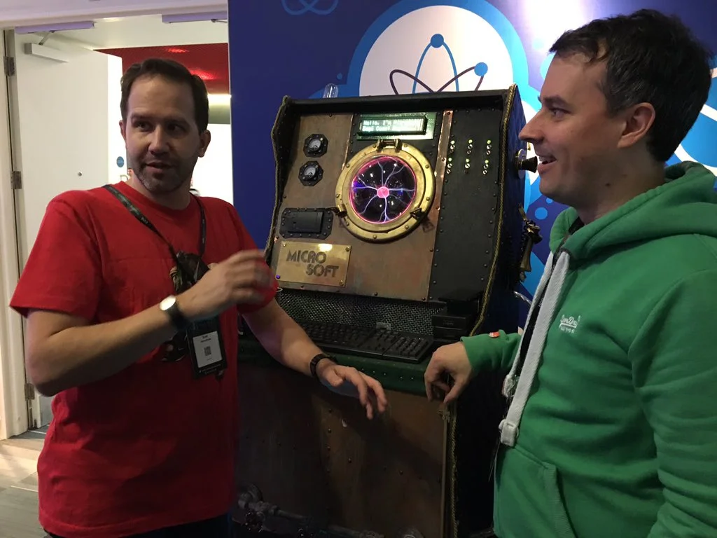 Admiral Edgecase on the Microsoft stand at NDC London, its plasma ball glowing in a brass porthole above a brass Microsoft plaque, with two people chatting beside it.
