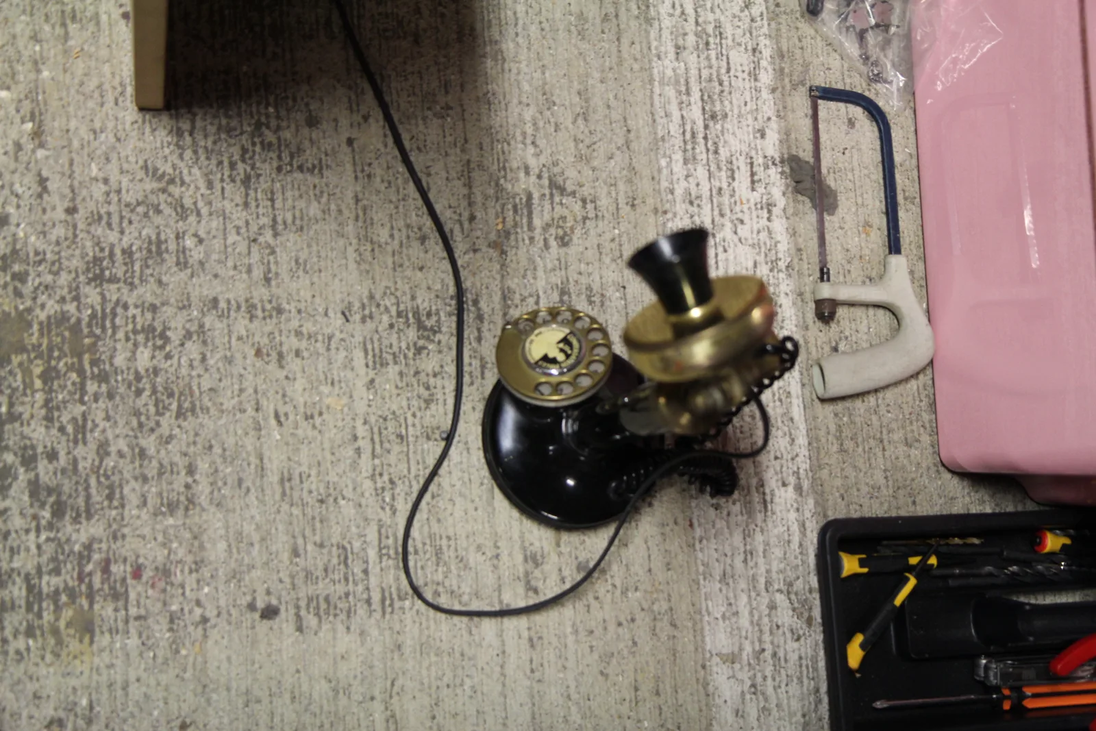 An old black candlestick telephone lying on the floor of the workshop next to a hacksaw.
